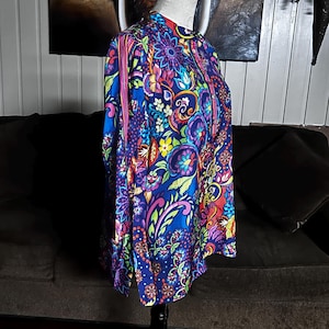 May include: A vibrant, long-sleeved shirt with a high collar, displayed on a mannequin. The shirt features a colorful floral pattern in shades of blue, purple, pink, yellow, and green. The design includes intricate floral and paisley motifs, creating a bold and eye-catching look.