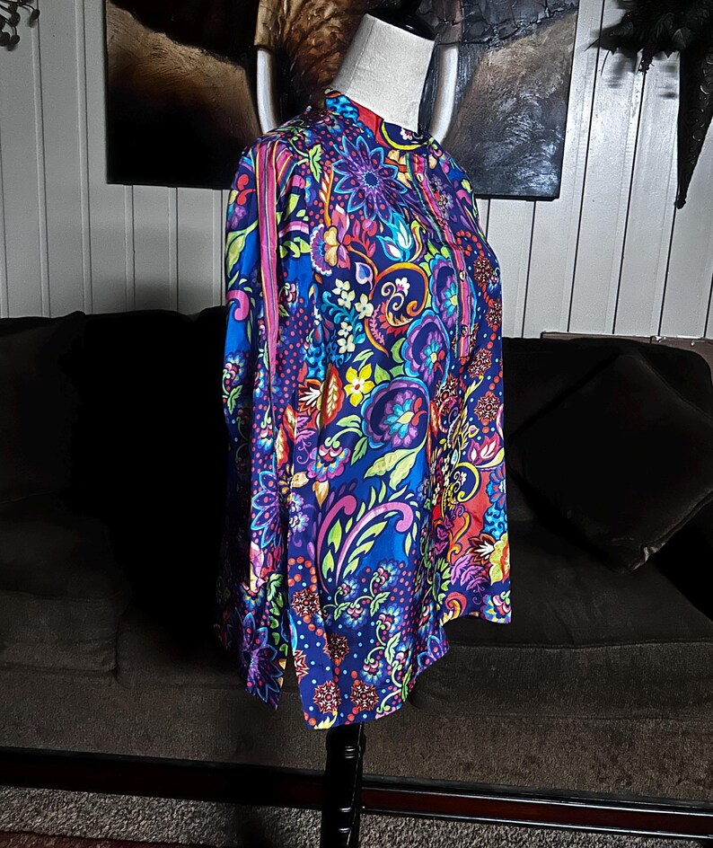 May include: A vibrant, long-sleeved shirt with a high collar, displayed on a mannequin. The shirt features a colorful floral pattern in shades of blue, purple, pink, yellow, and green. The design includes intricate floral and paisley motifs, creating a bold and eye-catching look.