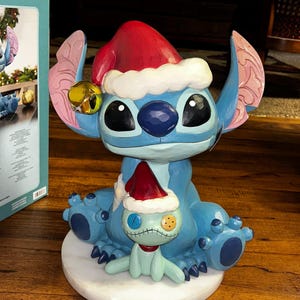 May include: A blue Stitch figurine wearing a red Santa hat with white trim and a gold bell. A smaller green doll with a Santa hat sits in front of Stitch. The figurine is on a white marble base.