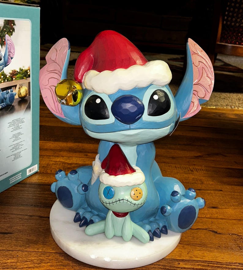 May include: A blue Stitch figurine wearing a red Santa hat with white trim and a gold bell. A smaller green doll with a Santa hat sits in front of Stitch. The figurine is on a white marble base.