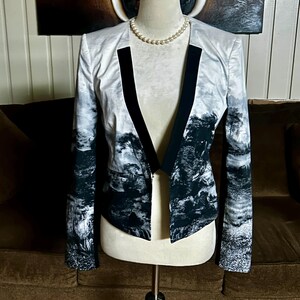 May include: A white and black landscape print blazer with black lapels. The jacket is displayed on a mannequin with a pearl necklace. The landscape design features trees and mountains.