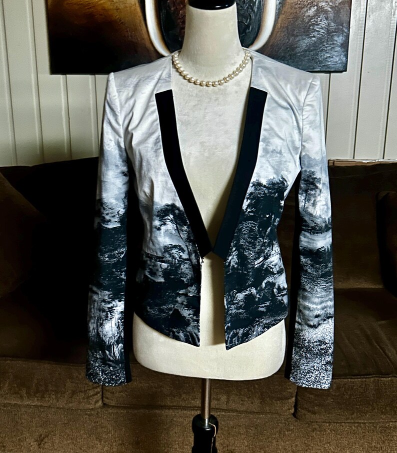 May include: A white and black landscape print blazer with black lapels. The jacket is displayed on a mannequin with a pearl necklace. The landscape design features trees and mountains.