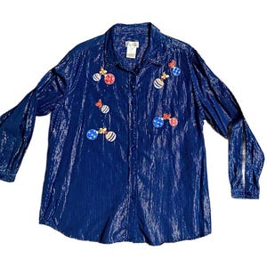May include: A long-sleeved, button-down shirt in a deep blue color with a subtle, shimmering vertical texture. The front of the shirt is decorated with several colorful, round Christmas ornament appliqués, each featuring a small gold-toned bow detail.