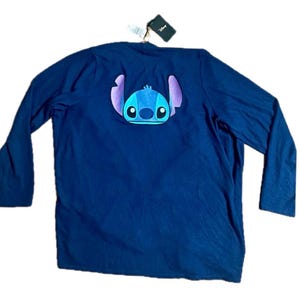 May include: Navy blue long-sleeve shirt featuring a Stitch graphic on the back. The graphic is a detailed, colorful depiction of the character's face with blue, purple, and white accents. The shirt has a tag attached.