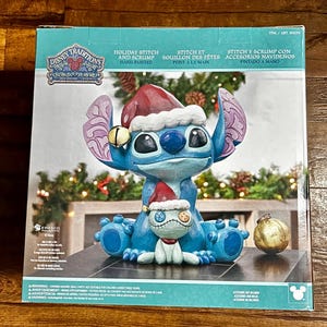 May include: A Disney Traditions holiday figurine featuring Stitch and Scrump in Santa hats. The blue Stitch figure has large ears and a bell. The figurine is hand-painted and includes a gold ornament.