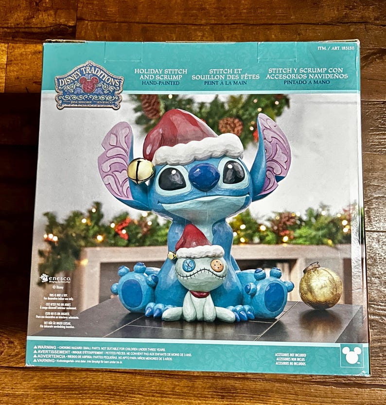May include: A Disney Traditions holiday figurine featuring Stitch and Scrump in Santa hats. The blue Stitch figure has large ears and a bell. The figurine is hand-painted and includes a gold ornament.