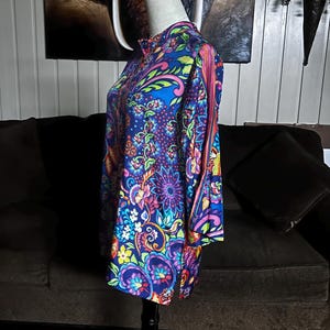 May include: A vibrant, long-sleeved tunic with a high collar, displayed on a mannequin. The tunic features a colorful floral and paisley pattern in shades of blue, purple, pink, and green. The fabric has a silky sheen, and the design is reminiscent of vintage styles.