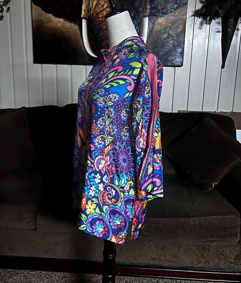 May include: A vibrant, long-sleeved tunic with a high collar, displayed on a mannequin. The tunic features a colorful floral and paisley pattern in shades of blue, purple, pink, and green. The fabric has a silky sheen, and the design is reminiscent of vintage styles.