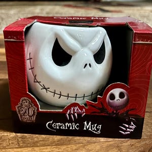 Puede incluir: Taza de cer&aacute;mica blanca con la cara de Jack Skellington, con detalles cosidos y ojos negros. La taza est&aacute; en una caja roja con las palabras "Ceramic Mug" y un logotipo del 20 aniversario.