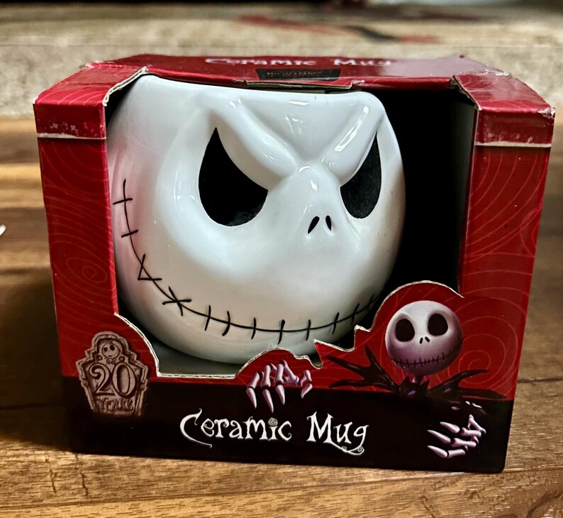 Puede incluir: Taza de cer&aacute;mica blanca con la cara de Jack Skellington, con detalles cosidos y ojos negros. La taza est&aacute; en una caja roja con las palabras "Ceramic Mug" y un logotipo del 20 aniversario.