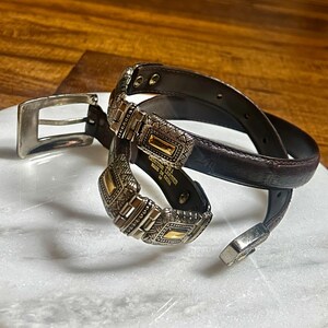 May include: A brown leather belt with silver and gold-toned metal accents. The belt features a decorative buckle and matching belt loop. The belt is coiled on a white surface.