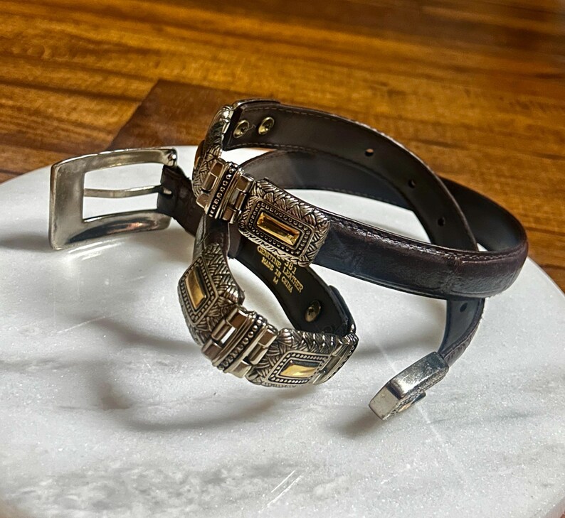 May include: A brown leather belt with silver and gold-toned metal accents. The belt features a decorative buckle and matching belt loop. The belt is coiled on a white surface.