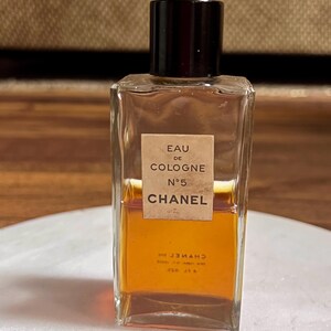 May include: A rectangular glass bottle of Chanel No. 5 Eau de Cologne. The bottle has a dark brown cap and a beige label with black text. The liquid inside is amber-colored. The bottle is on a white surface.