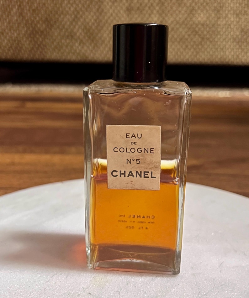 May include: A rectangular glass bottle of Chanel No. 5 Eau de Cologne. The bottle has a dark brown cap and a beige label with black text. The liquid inside is amber-colored. The bottle is on a white surface.
