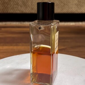 May include: A rectangular glass perfume bottle with a black cap. The bottle contains a golden-brown liquid. The label on the bottle reads "EAU DE COLOGNE N&deg;5 CHANEL". The bottle is on a white surface.