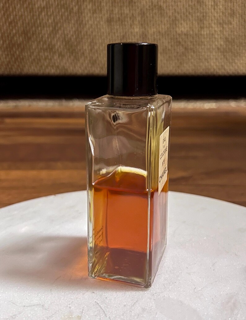 May include: A rectangular glass perfume bottle with a black cap. The bottle contains a golden-brown liquid. The label on the bottle reads "EAU DE COLOGNE N&deg;5 CHANEL". The bottle is on a white surface.