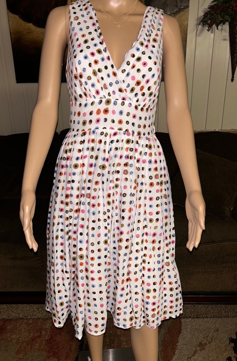 May include: White sleeveless dress with a V-neckline, adorned with a pattern of colorful polka dots in shades of pink, blue, brown, and black. The dress has a fitted bodice and a flared skirt, creating a classic silhouette.