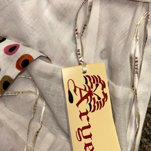 May include: White fabric with a colorful polka dot pattern. A tag with the brand name "Aryeh" is attached with a silver ribbon. The tag features a stylized design and the brand name in a decorative font. The fabric is folded.