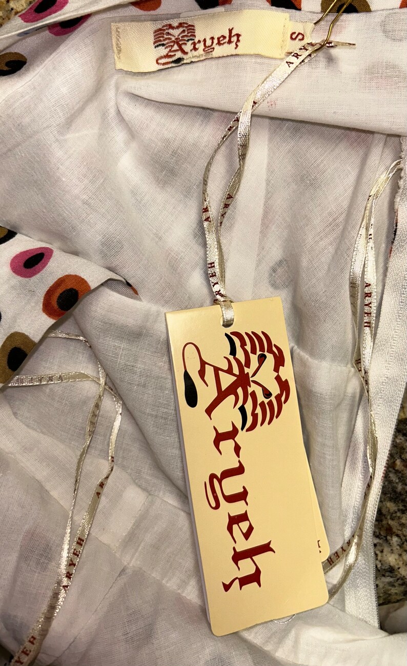 May include: White fabric with a colorful polka dot pattern. A tag with the brand name "Aryeh" is attached with a silver ribbon. The tag features a stylized design and the brand name in a decorative font. The fabric is folded.