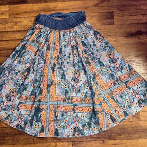 May include: A flowing skirt with a blue denim waistband and a vibrant, patterned design. The skirt features a mix of teal, orange, and purple hues in a geometric and floral print, creating a bohemian aesthetic.
