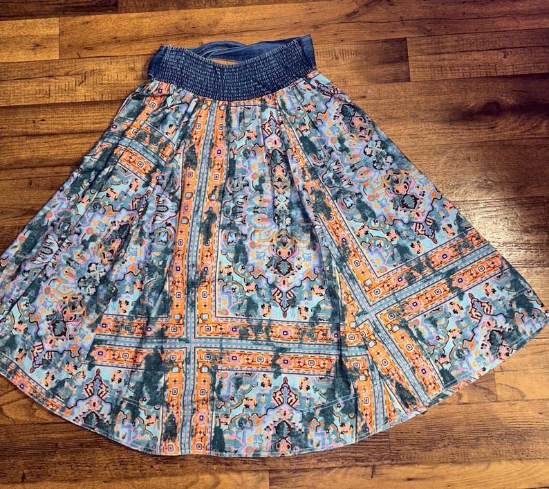 May include: A flowing skirt with a blue denim waistband and a vibrant, patterned design. The skirt features a mix of teal, orange, and purple hues in a geometric and floral print, creating a bohemian aesthetic.