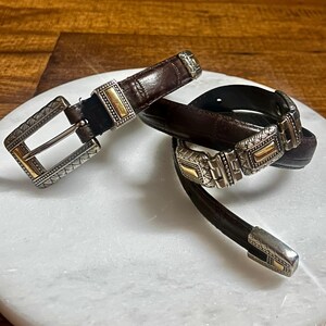 May include: Two brown leather belts with decorative silver and gold buckles. The belts are displayed on a white marble surface. The buckles have intricate designs and add a touch of elegance to the belts.