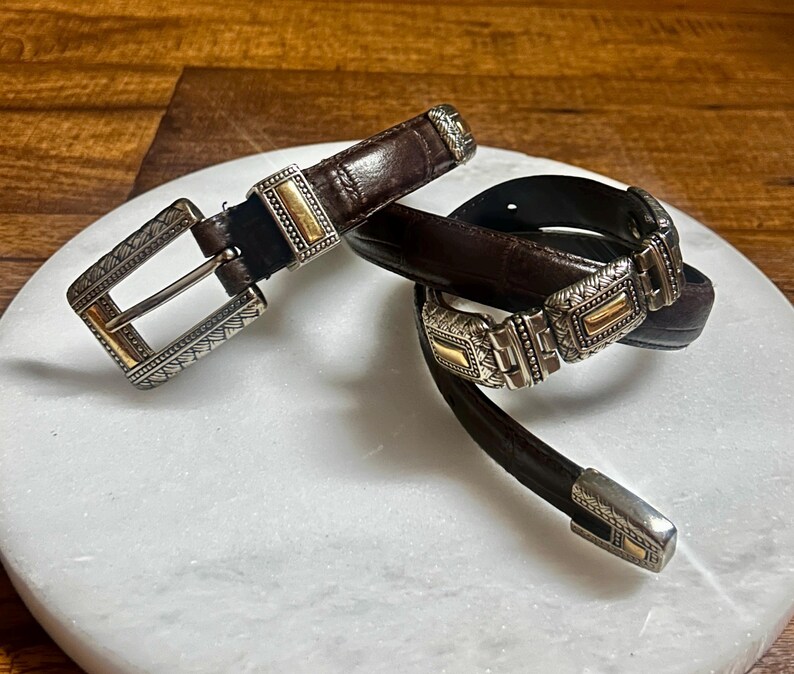 May include: Two brown leather belts with decorative silver and gold buckles. The belts are displayed on a white marble surface. The buckles have intricate designs and add a touch of elegance to the belts.