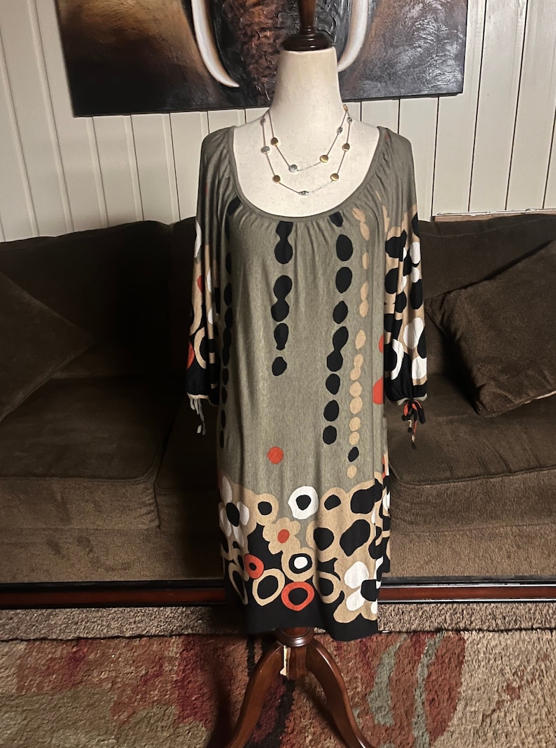 May include: A green, brown, black, white, and red patterned dress with a scoop neck and 3/4 sleeves. The dress is on a mannequin and has a beaded necklace around the neck.