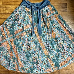 May include: A flowing skirt with a denim waistband and tie. The skirt features a colorful, patterned design in shades of teal, orange, and purple. The fabric has a soft, flowing drape, and the denim waistband adds a contrasting texture.