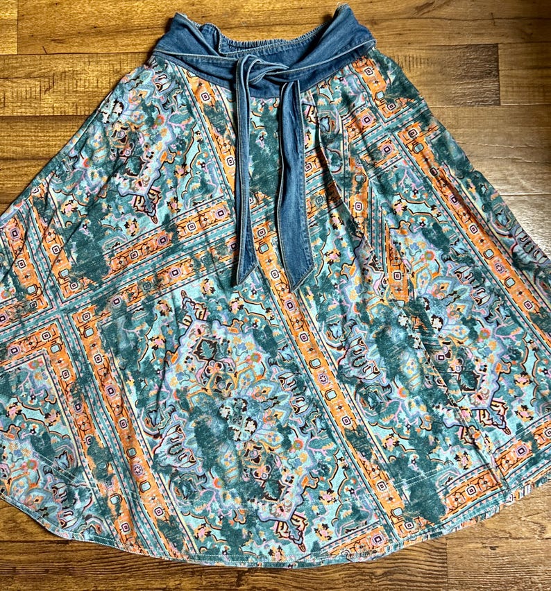May include: A flowing skirt with a denim waistband and tie. The skirt features a colorful, patterned design in shades of teal, orange, and purple. The fabric has a soft, flowing drape, and the denim waistband adds a contrasting texture.