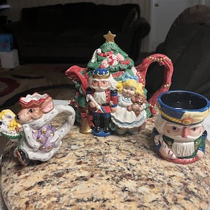 May include: A Christmas themed tea set with a teapot shaped like a Christmas tree with a nutcracker and a girl holding a teddy bear. There is also a small teacup shaped like a nutcracker and a creamer shaped like a mouse.