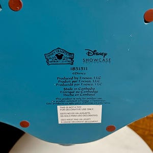 May include: A close-up of the bottom of a decorative Disney Showcase Collection item. The item is turquoise with red accents. Text includes "Disney Traditions", "Disney Showcase Collection", and "Made in Cambodia". A sticker states "This is not a toy, for decorative use only."
