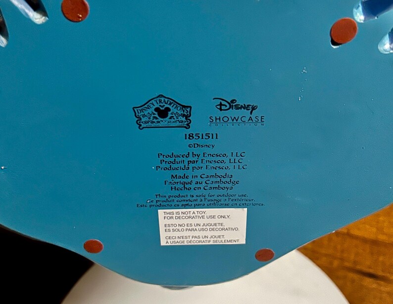 May include: A close-up of the bottom of a decorative Disney Showcase Collection item. The item is turquoise with red accents. Text includes "Disney Traditions", "Disney Showcase Collection", and "Made in Cambodia". A sticker states "This is not a toy, for decorative use only."