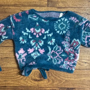 May include: A teal blue fuzzy sweater with a pink and green floral pattern. The sweater has a tie at the bottom.