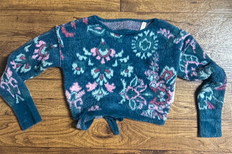 May include: A teal blue fuzzy sweater with a pink and green floral pattern. The sweater has a tie at the bottom.