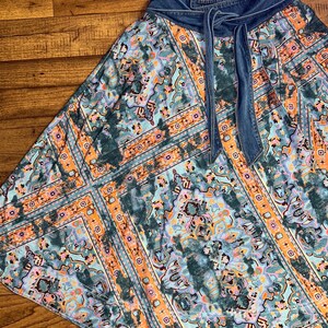 May include: A flowing skirt with a denim waistband and tie. The skirt features a colorful, patterned design in shades of teal, orange, and purple. The fabric has a soft, flowing drape, and the denim waistband adds a contrasting texture.