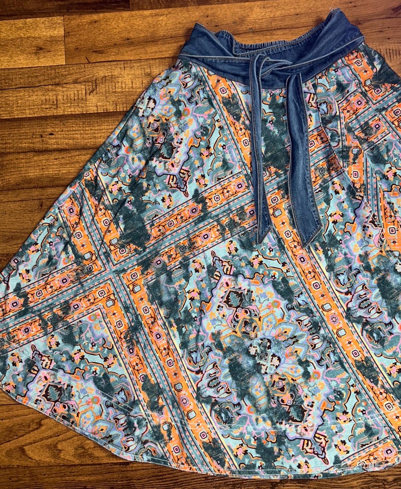 May include: A flowing skirt with a denim waistband and tie. The skirt features a colorful, patterned design in shades of teal, orange, and purple. The fabric has a soft, flowing drape, and the denim waistband adds a contrasting texture.