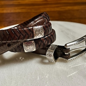 K&ouml;nnte beinhalten: Nahaufnahme von drei braunen Lederg&uuml;rteln mit silbernen Schnallen. Die G&uuml;rtel sind geflochten und haben dekorative silberne Akzente. Die Schnallen weisen aufwendige Designs auf. Die G&uuml;rtel sind auf einer wei&szlig;en Oberfl&auml;che aufgerollt.