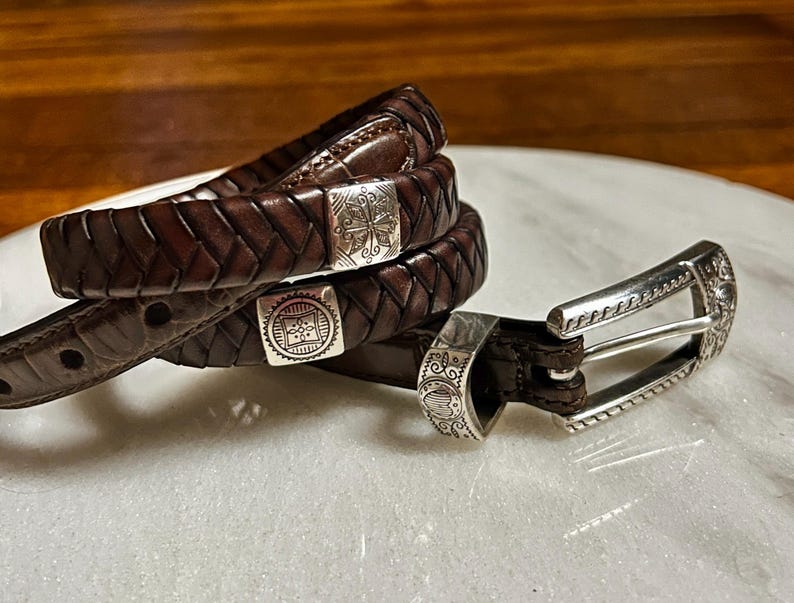 K&ouml;nnte beinhalten: Nahaufnahme von drei braunen Lederg&uuml;rteln mit silbernen Schnallen. Die G&uuml;rtel sind geflochten und haben dekorative silberne Akzente. Die Schnallen weisen aufwendige Designs auf. Die G&uuml;rtel sind auf einer wei&szlig;en Oberfl&auml;che aufgerollt.