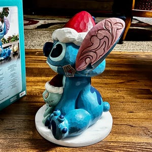 May include: A blue Stitch figurine wearing a Santa hat and holding a small plush toy. The figurine is on a white circular base. The ear has a pink and brown design. The figurine is a Disney collectible.