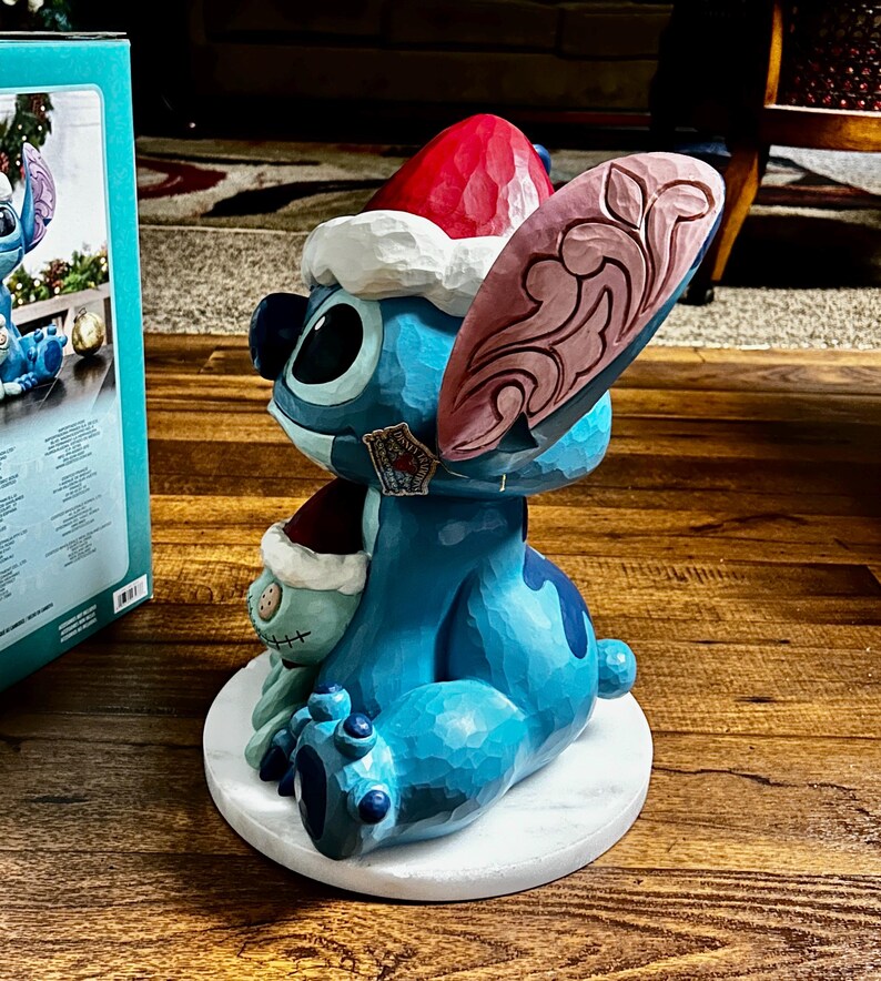 May include: A blue Stitch figurine wearing a Santa hat and holding a small plush toy. The figurine is on a white circular base. The ear has a pink and brown design. The figurine is a Disney collectible.