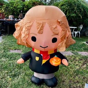 May include: An inflatable Halloween decoration in the shape of a cartoon character with brown hair, black robes, and a red and gold scarf. The character is holding an orange jack-o'-lantern. The decoration is standing on green grass.