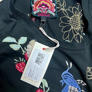 Puede incluir: Un vestido negro con bordados coloridos de flores y p&aacute;jaros. La prenda incluye una etiqueta de la marca Johnny Was Los Angeles en el cuello y una etiqueta de venta blanca sujeta con un cordel, que muestra el texto Johnny Was, Style J37325-4, Asymmetric Ruffle Flounce, Size L, Color BLK y johnnywas.com.