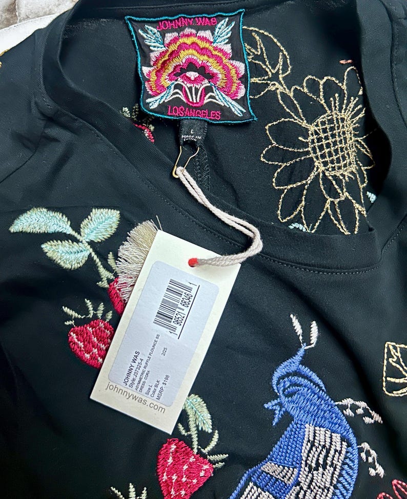 Puede incluir: Un vestido negro con bordados coloridos de flores y p&aacute;jaros. La prenda incluye una etiqueta de la marca Johnny Was Los Angeles en el cuello y una etiqueta de venta blanca sujeta con un cordel, que muestra el texto Johnny Was, Style J37325-4, Asymmetric Ruffle Flounce, Size L, Color BLK y johnnywas.com.