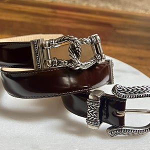 Puede incluir: Un cintur&oacute;n de cuero marr&oacute;n con una hebilla plateada con un dise&ntilde;o trenzado. La hebilla es adornada y tiene un dise&ntilde;o detallado.