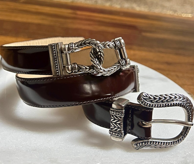Puede incluir: Un cintur&oacute;n de cuero marr&oacute;n con una hebilla plateada con un dise&ntilde;o trenzado. La hebilla es adornada y tiene un dise&ntilde;o detallado.