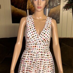 May include: White sleeveless dress with a V-neck and a fitted waist. The dress is adorned with a pattern of colorful polka dots in shades of pink, blue, brown, and gold. A gold necklace is visible.