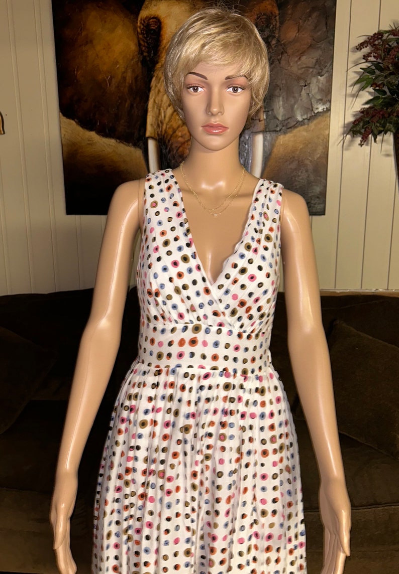May include: White sleeveless dress with a V-neck and a fitted waist. The dress is adorned with a pattern of colorful polka dots in shades of pink, blue, brown, and gold. A gold necklace is visible.