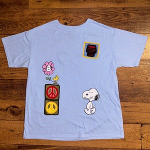 May include: Light blue t-shirt with embroidered patches featuring a flower, a peace sign, a cartoon bird, and a cartoon dog. The patches are yellow, red, green, and black.