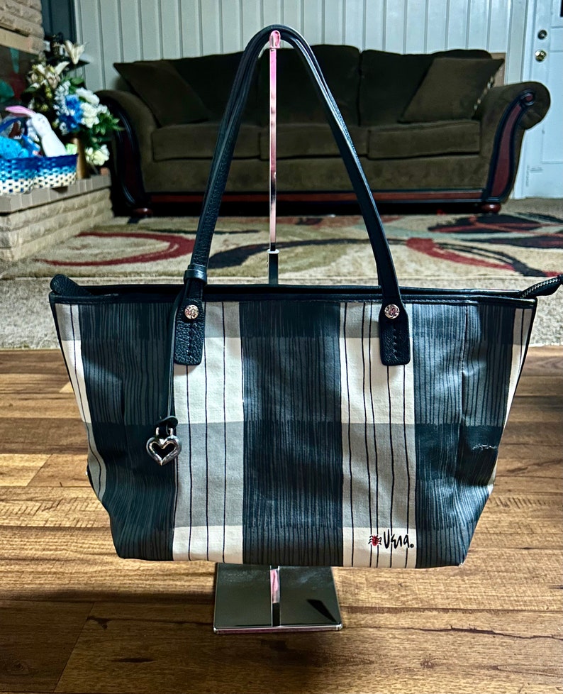May include: A black and white plaid tote bag with black leather handles and a silver heart charm. The bag has the word "Vera" embroidered on the bottom in red.